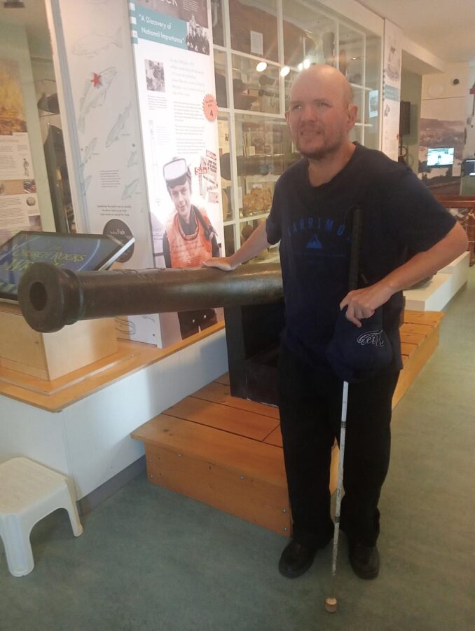 Tony touching an old cannon inside Teignmouth and Shaldon Museum (Teign Heritage Centre). The cannon is part of a display about the Church Rocks Wreck in the Sea Gallery.  In 1975, the first of six bronze cannons was found by Simon Burton, a local 13 year old, while snorkelling in the area. This cannon was a medium-sized ‘saker’ cannon bearing the initials of the Venetian gunmaker Sigmund Alberghetti. Two further guns (‘minions’) and three swivel guns were subsequently found, all in remarkable condition. The wreck the cannons came from dated from the  16th century and was found 200 yards off the beach at Teignmouth opposite St Michael’s Church. All the artifacts found are on display in the museum. At present significant parts of the ship remain on the seabed, buried under the sand. An information board behind includes a large photo of the boy who found the cannon wearing a life jacket and snorkelling gear.