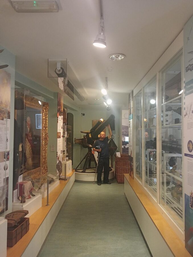 Looking along a narrow gallery with display cases down the sides. At the far end Tony is touching a medium-sized gun mounted on a brass stand that allows it to be quickly swivelled round towards a target. This is the Peronne Gun which was recovered from a wreck six miles off Teignmouth in 1993. It belonged to a merchant ship called the Peronne which was torpedoed and sank during World War One on 1st September 1917. At that time ships were obliged to carry defensive armoury and travel in convoy to avoid the German submarines. The one and a half ton gun has been restored and loaned to the museum.