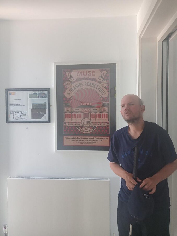 Tony in the upstairs room of Teignmouth and Shaldon Museum. He is next to a framed poster advertising two concerts by the rock band Muse that took place at Teignmouth Pier on 4th and 5th September 2009. The poster is titled "A Seaside Rendezvous" and has an historic stylised design depicting the pavilion on Teignmouth Grand Pier.  Muse formed in Teignmouth in 1994. There is also a smaller frame on the wall that contains photos and a signed concert ticket likely from one of the same concerts.