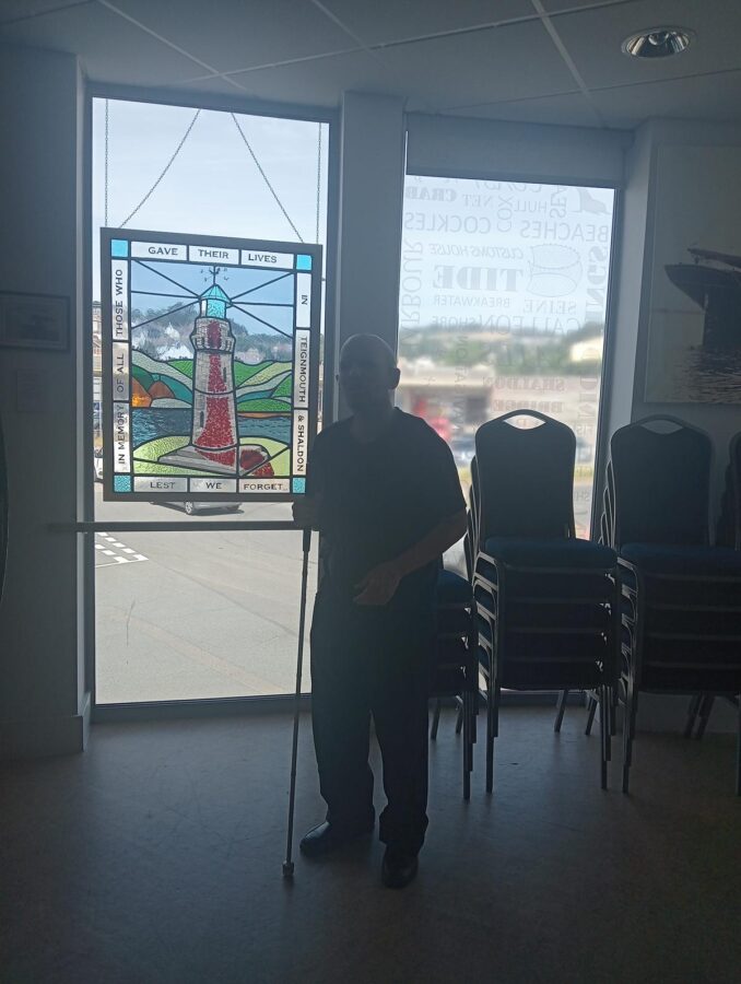 Tony in the same room as before alongside a colourful stained-glass panel hanging in a window. The panel depicts a red and white lighthouse with the sea, hills and blue sky in the background. Words around the edge read: "In memory of all those who gave their lives in Teignmouth & Shaldon lest we forget". This could be memorial to locals who died in the World Wars or one to lifeboat personnel. The room is part of the museum, but is also used as a community and event space.