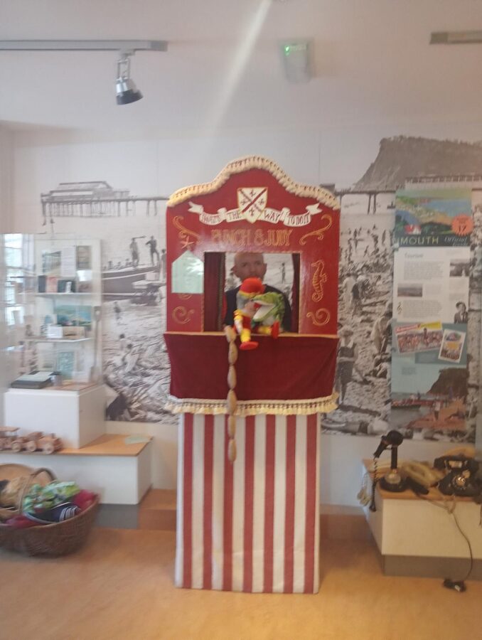 A traditional Punch and Judy puppet theatre booth at Teignmouth and Shaldon Museum (Teign Heritage Centre). The booth is decorated in red and white stripes with gold trim, and the top has a sign that reads "Punch & Judy", along with a crest that includes the words "That's the way to do it!" Tony is stood behind the booth,  visible through the opening, and he is holding two puppets: one appears to be Punch and the other is the crocodile. A long string of sausages are dangling from the front of the booth: this is a classic prop in Punch and Judy shows.