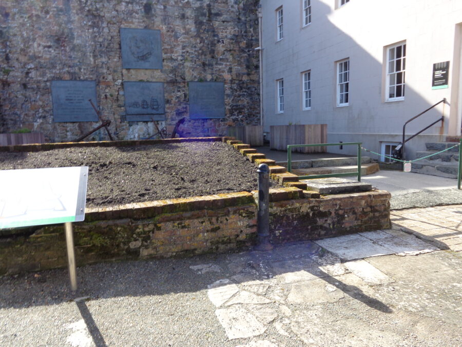 A stone and gravel path at the entrance to Castle Cornet in St. Peter Port, Guernsey. In front is a stone wall with four large bronze plaques attached. One of the plaques depicts a line of sailing ships. The writing on the other plaques is too distant to read. At the foot of the wall are three metal ship's anchors. The cream coloured building on the right is the Lower Barracks which dates from 1745 to 1750. 