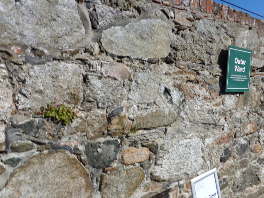 The Outer Ward at Castle Cornet. A stone outer wall runs down the left side of the photo. This courtyard would have originally been used for exercise and parades. In the background a higher stone and brick wall rises up with fortified buildings beyond. 