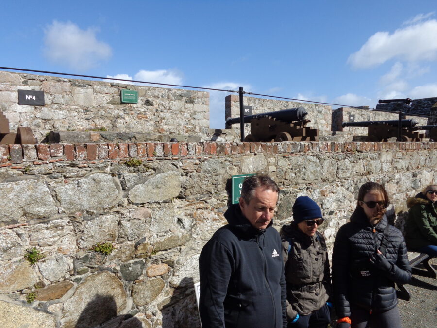 View of the Saluting Battery at Castle Cornet. A row of old cannons on metal carriages are pointing through gaps in the battlements. The cannons in view are numbered 2 and 3. Other visitors can be seen standing and sitting in the foreground. 