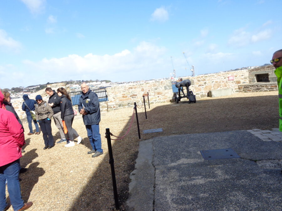 A group of visitors in a gravel covered area at Castle Cornet. The stone outer battlements can be seen around the edge with a cannon mounted on a carriage pointing outward through a gap in the wall. Parts of St. Peter Port are visible across the harbour in the middle distance.