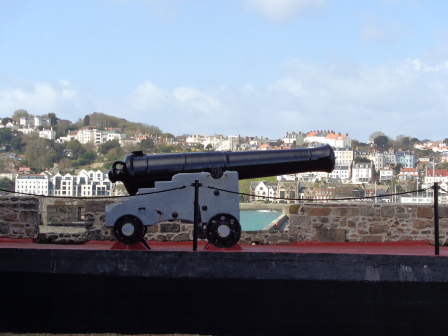 Another old cannon on a carriage. It is on a walkway on the battlements of Castle Cornet. There is a scenic view of St. Peter Port in the background with many white painted buildings on the hillside rising up from the sea front and harbour.