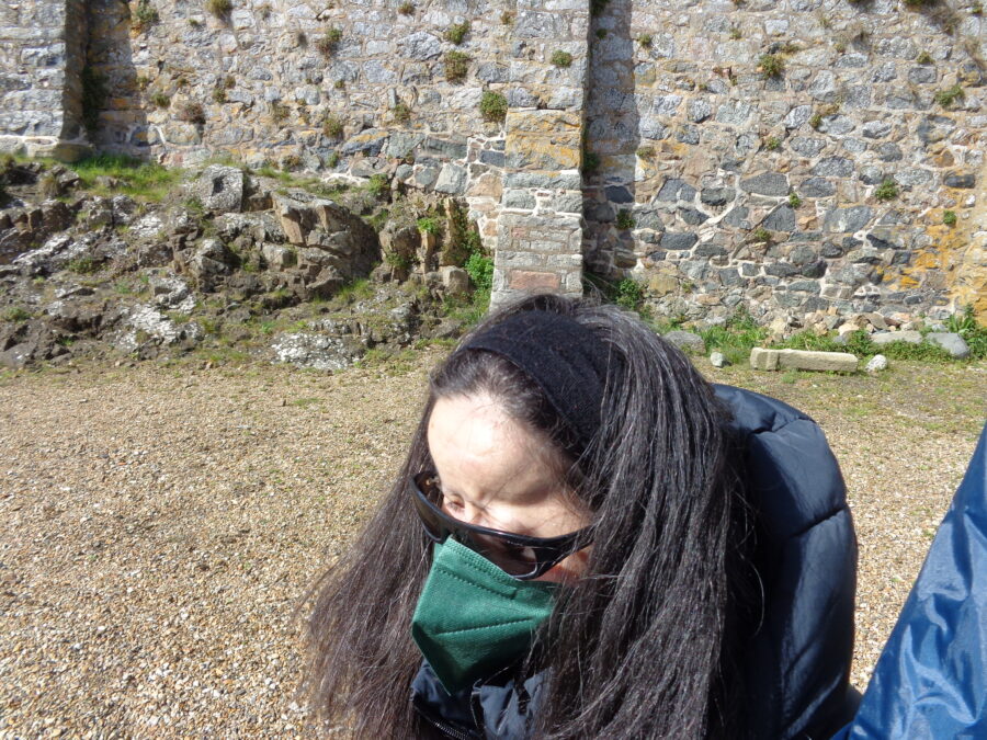 Photo of Tatiana standing in one of the courtyards at Castle Cornet. She is wearing a black headband, dark sunglasses, a green face mask and a warm blue jacket.