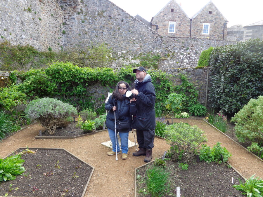 Tony and Tatiana stood together in the formally laid-out Lambert's Garden at Castle Cornet. Various stone walls and buildings are visible in the background.