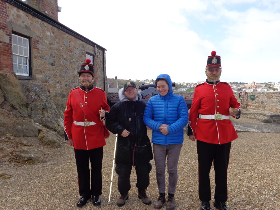 Four people stood in the castle's main courtyard. The two men at the ends of the group are formally dressed in bright red military-style tunics with black trousers and white belts. They are wearing black hats with red bands. The hats have silver badges attached to the front and red pommels on top. These men are known as Castle Keepers: museum staff who wear replica Guernsey Militia uniforms and are involved with the firing of the "Noon day Gun". The two people in the centre of the group are Tony and a woman who could be a visitor or a member of staff. The courtyard is surrounded by low stone walls and a building to the left. There is gravel under foot. Buildings of St. Peter Port are visible in the distance.