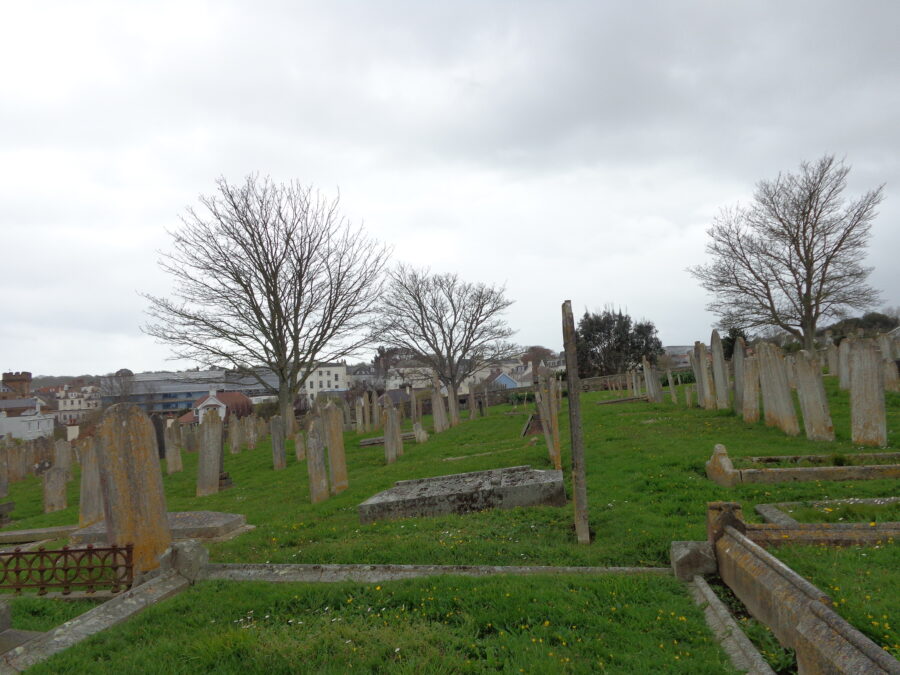 Rows of weathered-looking gravestones at Candie Cemetery. Some of the gravestones are leaning or tilted and a few are surrounded by low stone borders. Leafless trees stand among the graves, and beyond the cemetery, buildings of St. Peter Port can be seen, many painted white or cream.