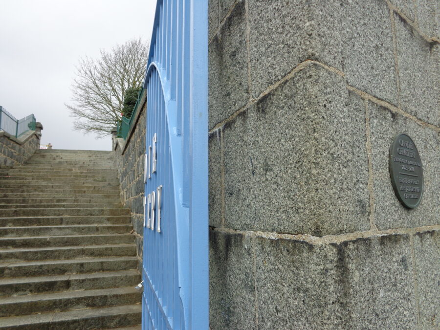Blue-painted metal gates at the entrance to Candie Cemetery. Beyond a long flight of around 50 steps leads up into the cemetery. A metal plaque to the right remembers the replacement of the cemetery gates in 2005-2006.