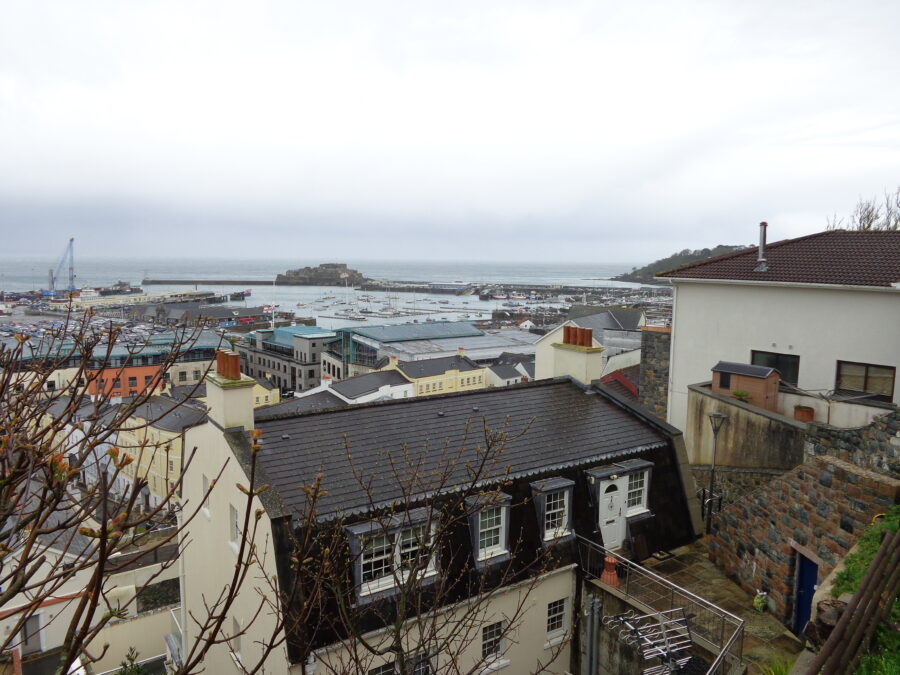 Good view over the rooftops of St. Peter Port town towards the harbour. Lots of boats, mostly quite small, can be seen in the harbour. Two harbour cranes are also in view. At the far side of the harbour, Castle Cornet is visible, attached to the mainland by the harbour wall. 