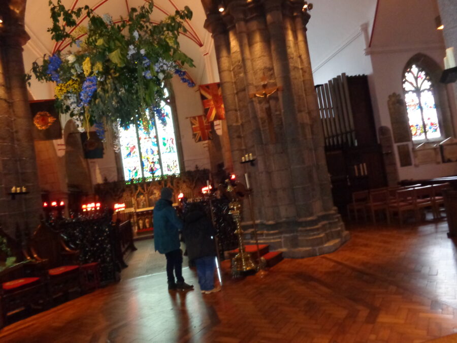 Interior of the Town Church, the parish church of St Peter Port. Tatiana can be seen standing with a tour guide in front of a brass lectern in the form of an eagle. The photo is looking towards the altar where rows of candles have been lit. There are stained-glass windows behind. A large stone column and an organ can also be seen. There is wooden  parquet flooring. This church was completed in its current form by 1466, but a small chapel is known to have existed here in 1020. As well as being the parish church of St. Peter Port this is also considered to be Guernsey's main church.
