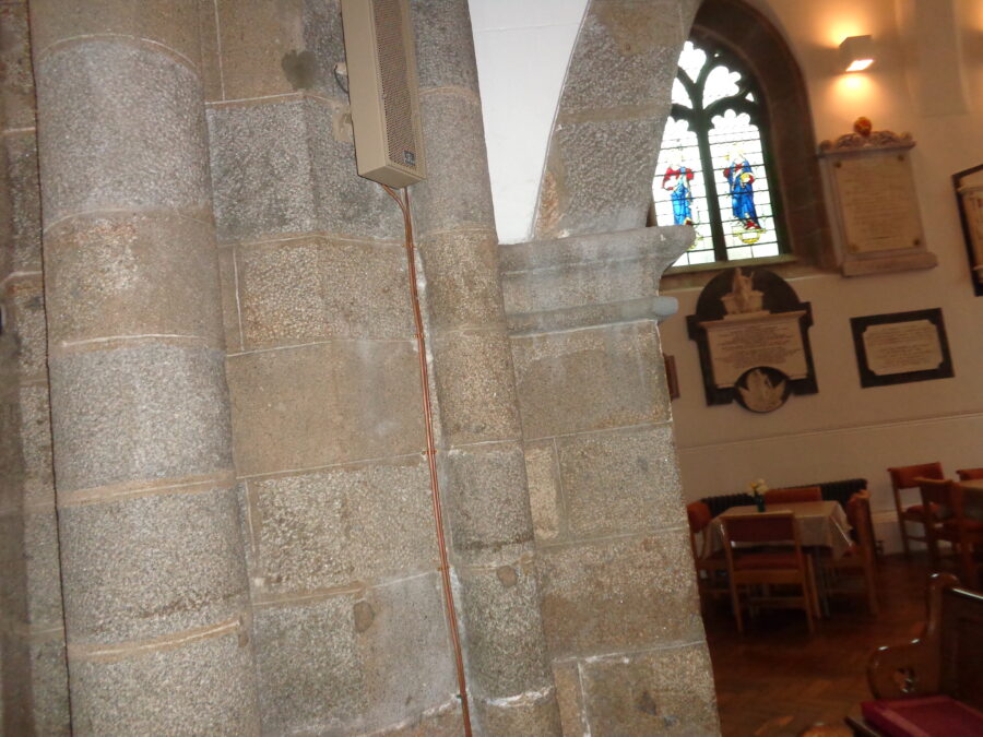 A stone pillar and archway inside the Town Church. To the right a stained-glass window can be seen with several memorial plaques attached to the wall beneath. There are chairs, tables and pews in front. 