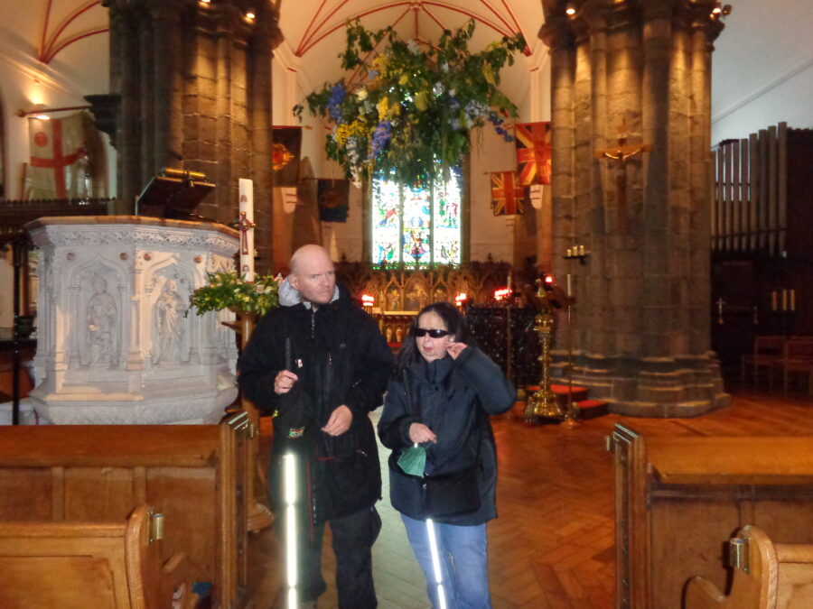 Tony and Tatiana near to the stone pulpit decorated with carved figures in Town Church. Greenery and flowers are hanging from above. The altar with candles lit is visible in the background. Various flags are hanging from columns inside the church: two are Union Jacks with an extra emblem in the centre.