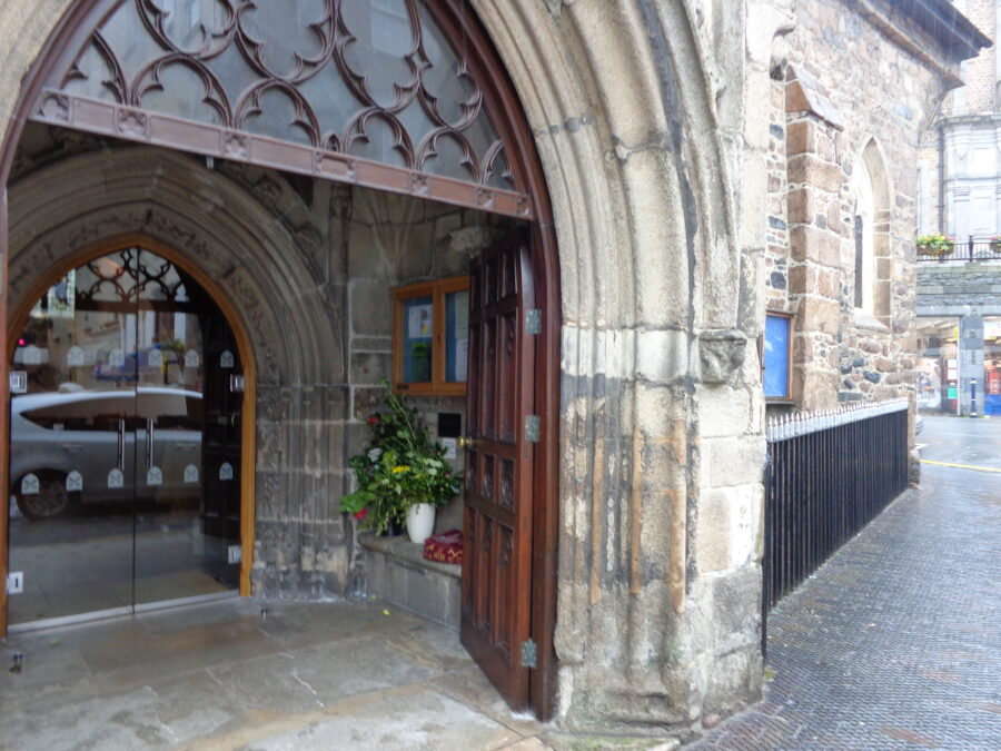 The main entrance to Town Church. There is a stone archway with large heavy wooden doors. Within there is a porch with a second set of glass doors beyond. There is a vase of flowers and a noticeboard inside the porch. The old stone-exterior-walls of the church can be seen.