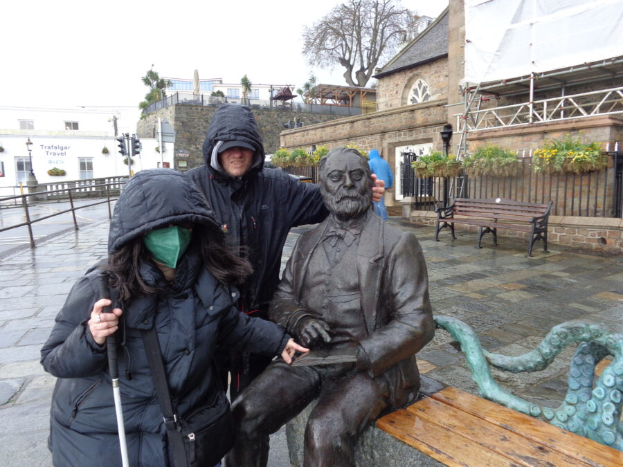 Tony and Tatiana wrapped up against the cold and wet evening weather, beside a bronze statue of 19th century writer, Victor Hugo, who is sat on a bench. Victor Hugo is depicted with a beard, wearing a suit and bow tie, and holding a book. The tentacles of a sculpted octopus curl around the back of the bench: this is a character from one of Hugo's novels. Tony and Tatiana are touching the statue. 