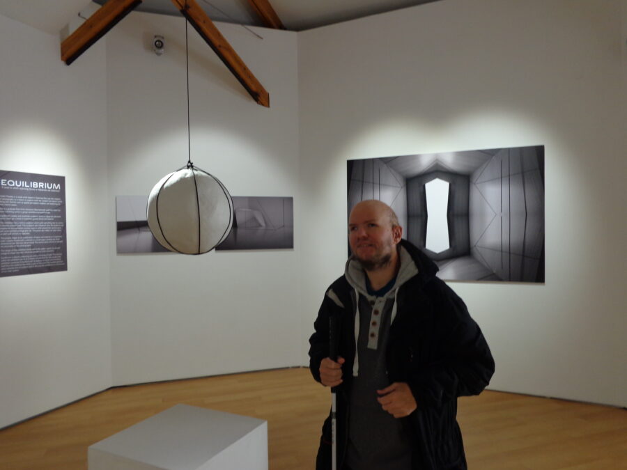 Tony in a room inside the Guernsey Museum and Art Gallery. A white sphere is hanging from the ceiling supported by black cord. Abstract monochrome artworks are displayed on the walls. There is an information board titled "Equilibrium", which might be the title of the collection of artworks on display. 