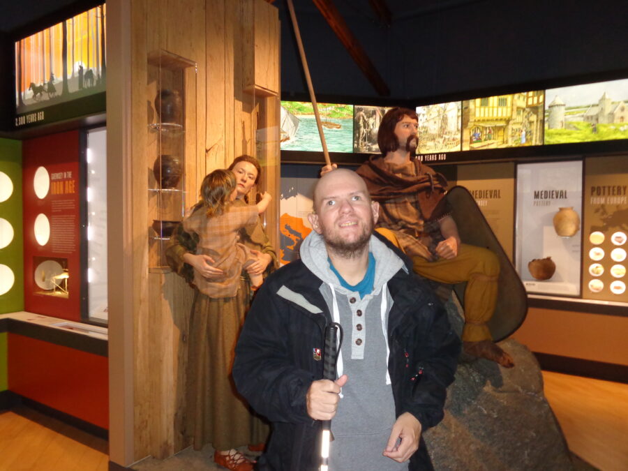 Displays about Guernsey at different points in its history at Guernsey Museum and Art Gallery. To the left is a display about Iron Age Guernsey and on the right a room dedicated to Medieval Guernsey. In the foreground are mannikins in Iron Age dress. There is a man holding a spear and shield. He is wearing a brown cloak. Next to him is a woman holding a child. She is wearing a dress made of coarse yellow-brown cloth.