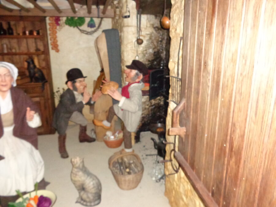 Re-creation of a historic kitchen at Guernsey Museum and Art Gallery. There is a mannikin of a woman in a bonnet and apron on the left. Two male mannikins appear to be talking to the right. One of the men is holding a loaf of bread. One man is wearing a top hat and the other a cap. There is a fireplace to the right and a dresser at the back of the room. There are two cats: one sitting on the floor and the other on the dresser. This is perhaps a scene from the 18th or 19th centuries.