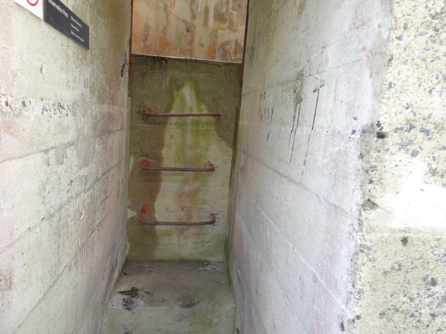 Entrance to the German Naval Signals Headquarters in St. Peter Port. There is a short concrete passageway with three metal rungs embedded in the far wall. A modern sign says Observation Post. The rough concrete walls are stained with algae. This was the headquarters for the German Naval Commander of the Channel Islands during the Second World War as well as the main communications centre. All the important radio signals traffic for the German Forces in the Channel Islands was handled here. The Channel Islands Occupation Society has refitted one of the bunkers as a museum. It is situated in St. Jacques, St. Peter Port, next door to La Collinette Hotel.