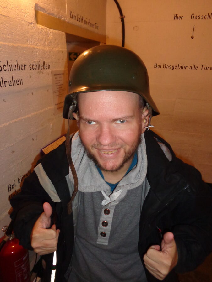 Tony wearing a green German military helmet, smiling and giving two thumbs up. There are again white painted concrete walls with instructions in German written on them.