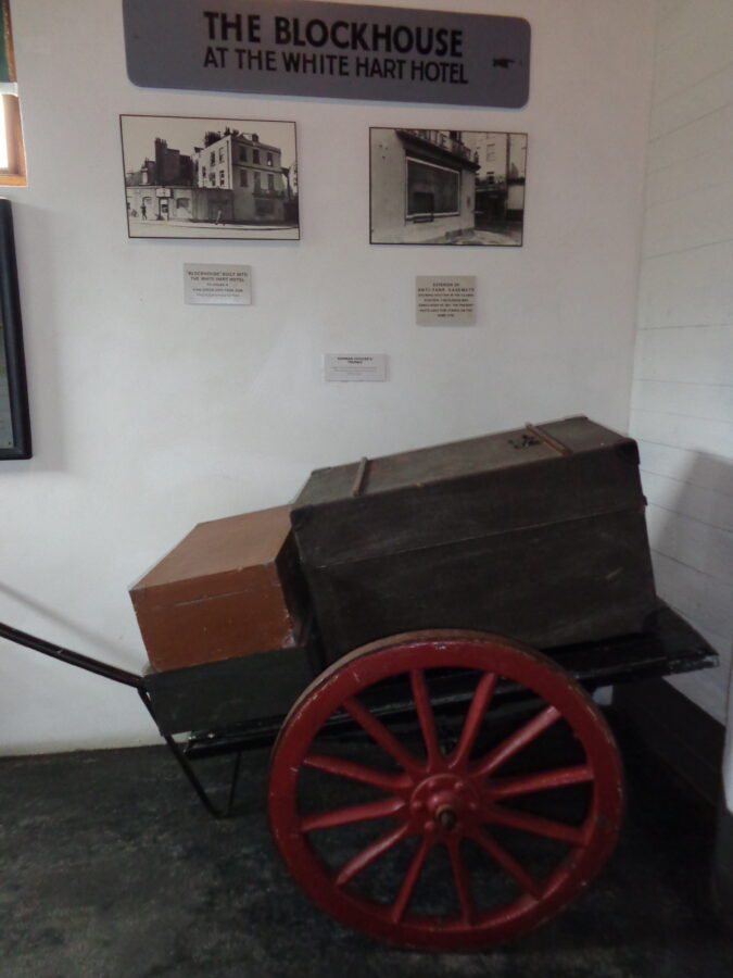 A pair of German Officers' trunks (suitcases) displayed on a wooden hand cart. There are also two black and white photos from 1944 showing a "blockhouse" built into the White Hart Hotel, St. Peter Port. This was a defensive structure to house an anti-aircraft gun.
