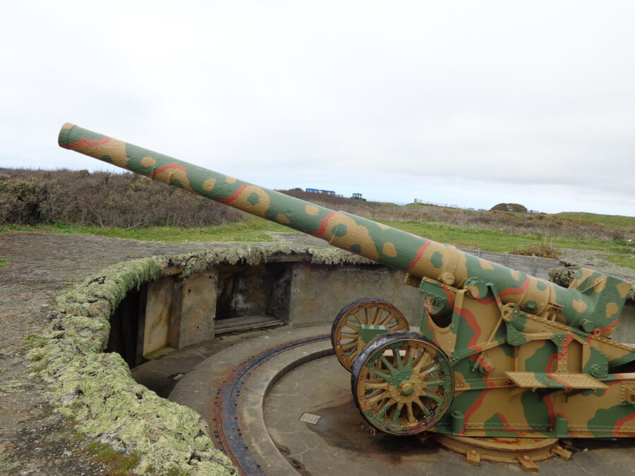 A Germany artillery gun painted in camouflage colours and situated in a shallow circular gun pit. It is constructed so that the gun can quickly be rotated to fire in different directions. It is located at Batterie Dollman on the south-west coast of Guernsey. The restored gun pit is the last of its type in Europe. The 10 tonne, 22cm gun was manufactured in France and was restored by Guernsey Armouries in 1997.