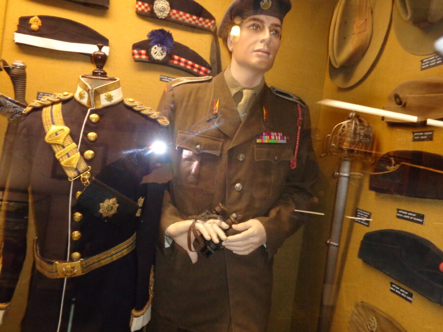 A collection of British military caps. There is also a mannikin of a British soldier in uniform. He is wearing a khaki brown jacket and a blue cap, and is holding a pair of binoculars. A smarter formal military uniform with gold buttons is displayed along side. 