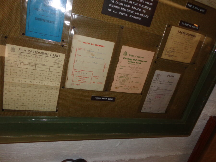 A display case containing a collection of ration books from the period of German occupation between 1940 and 1945. They include a Fishing Ration Card and a Clothes and Footwear Ration Book both issued by the States of Guernsey. The fronts of these documents typically include the name and address of the person it was issued to and the issue date. There is also a day permit to access the harbour, which unlike the other documents, is in German and has a German military stamp.