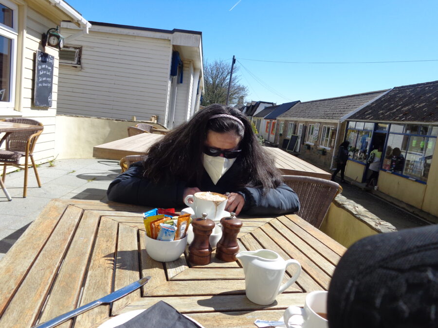 Tatiana enjoying a cup of coffee at a cafe on The Avenue, which is the main street in Sark. The photo shows small single storey buildings many housing shops and other businesses.