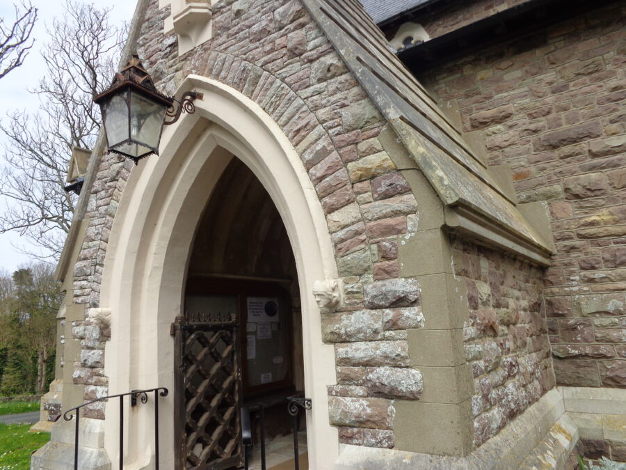 The entrance to St. Anne's church on Alderney's main street (Victoria Street). There is a stone porch with a Gothic-arched entrance gate. A metal lantern hangs above the arch. The church is dedicated to St. Anne, the mother of the Virgin Mary. The present church was designed by renowned Victorian architect George Gilbert Scott and was consecrated in 1850. This partially replaced an older church dating back to the 12th or 13th centuries. The clock tower of the old church was retained. The building is surrounded by a churchyard, which dates back to at least the 7th century, and a chapel is thought to have existed here from the same date.