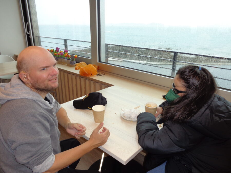 Tony and Tatiana sitting in a cafe. Through the window there is a view of the bathing pools and the sea beyond. It is raining outside making the view a little hazy.