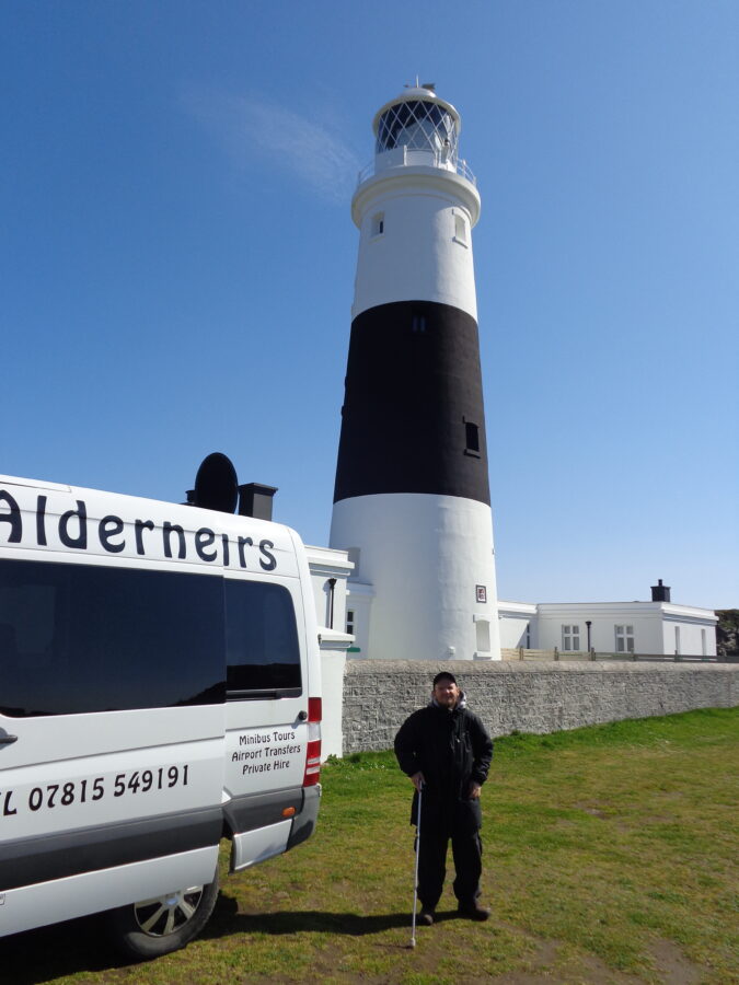 Alderney Lighthouse (also known as Mannez Lighthouse) located at Quesnard Point on the North-East coast of the island. The conical granite lighthouse is 32 metres (105 ft) in height. It was constructed in 1912 to protect shipping from the dangerous waters of the Alderney Race and the numerous rocks surrounding Alderney. It was electrified in 1976, and automated in 1997, when the last resident lighthouse keeper left. The lighthouse is painted black and white. Tony is standing in the foreground next to a minibus.