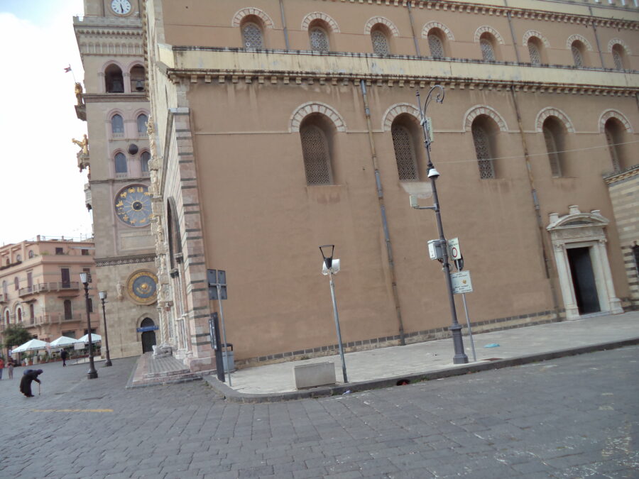 Looking towards one of the side walls belonging to Messina's historic cathedral. The front façade can also be seen at a very acute angle as well as one side of the bell tower. The side wall is relatively plain. It is covered with tan coloured plaster and there are simple arched windows. There is a more elaborate side doorway surrounded by neoclassical decoration. The distinctive bell tower claims to contain the most complex astronomical clock in the world and is one of the city's main attractions. It was constructed by the Ungerer Company of Strasbourg in 1933. The side of the tower that is in view contains three mechanical features, which can distantly be seen. The dial near the bottom is a perpetual calendar that shows the full date and the time of moveable Christian feasts. Further up, another large round dial with the details highlighted in gold, is a planetarium. This shows the position of the nine planets with the signs of the zodiac around the edge. Further up still is a 1.2 metre sphere showing the phase of the moon. The sphere is half-gold and half-black. In the photo, the visible part is almost all black, with just a thin sliver of gold down the left side. This indicates there was a new moon when this photo was taken. The cobbled square surrounding the cathedral can also be seen.