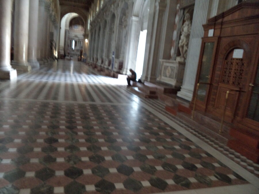 Inside Messina Cathedral looking down one of the side aisles. The floor is decorated with geometric marble tiles. There are large marble columns running down the left side. The wall down the right side contains elaborate neoclassical decoration including statues. A wooden structure with two doors can be seen immediately to the right. This is a confessional booth.