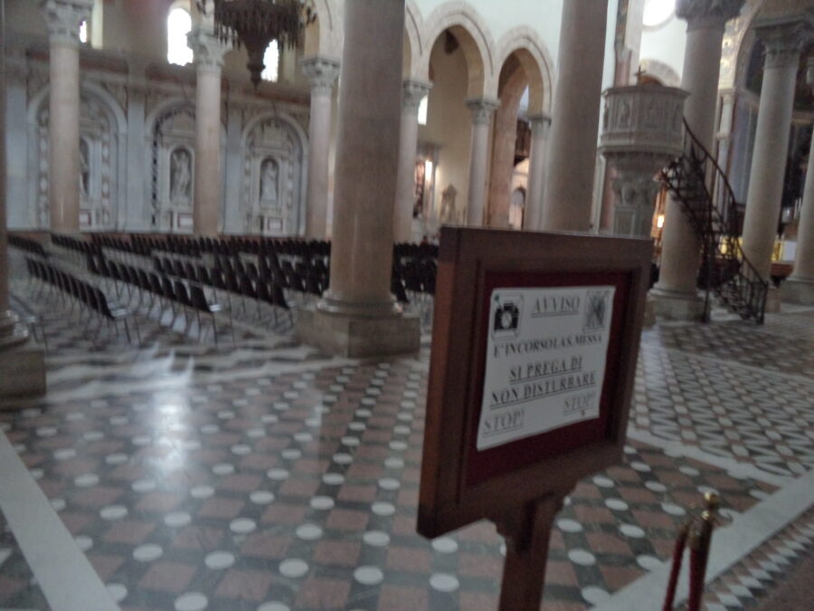 Interior of Messina Cathedral. The photo looks across the central part of the nave. There are rows of empty chairs. Large reddish marble columns supporting Norman arches run down both sides. To the right there is a high stone pulpit accessed using a spiral metal staircase.