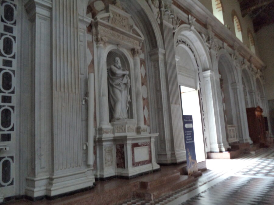 Looking along the inside of the entrance wall within Messina Cathedral which is adorned with neoclassical decoration. The large arched main entrance doorway can be seen in the centre. Closer to the camera, a marble statue stands in an alcove.  The statue is probably a saint and depicts a large bearded man dressed in a robe.