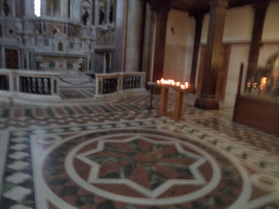 Slightly blurry view towards a large elaborately decorated marble side altar inside Messina Cathedral. Marble balustrades form a barrier at the front of the altar. The floor in front is covered with geometric marble tiles in white, black and red, including a large 8-pointed star. Candles are lit on tables on the right side.