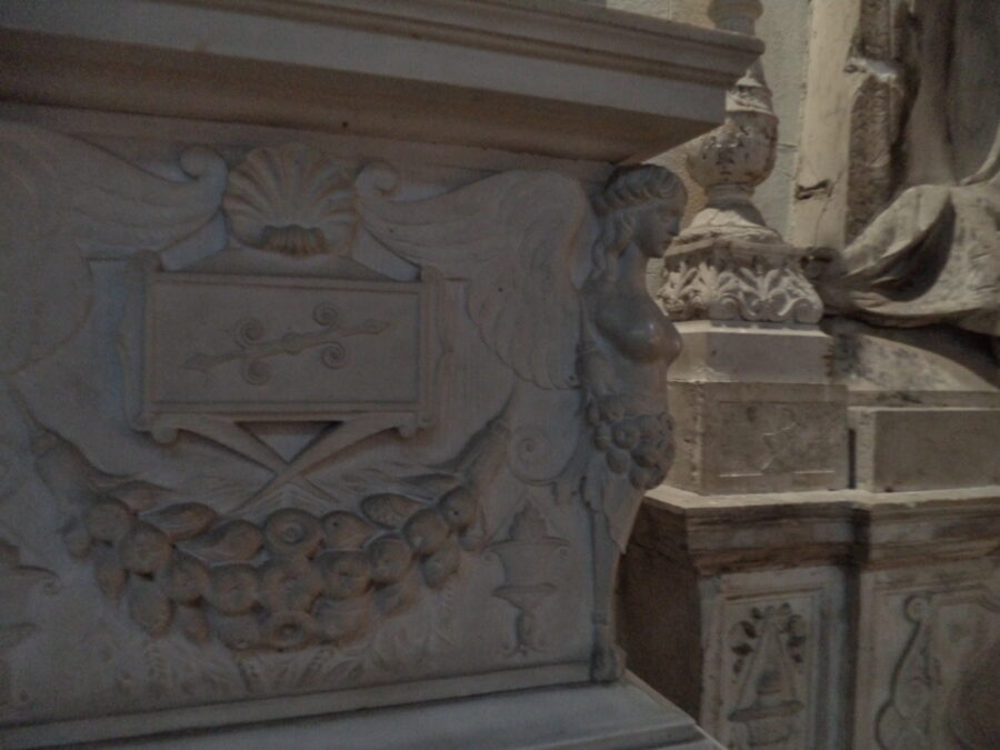 A large carved stone tomb inside Messina Cathedral. The tomb appears to have a nautical theme, which includes a shell motif in the centre, and a bare breasted female figure projecting from one corner. The female figure has wings, so is probably an angel, but also resembles a mermaid.