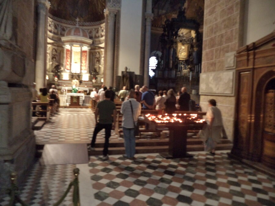 A slightly blurred view of a service taking place in front of a side altar within Messina Cathedral. The photo is taken from the back with an aisle running down the centre and pews at either side half full with people standing. A priest dressed in green can indistinctly be seen standing at the altar. There is also a large stand filled with lit candles at the back.