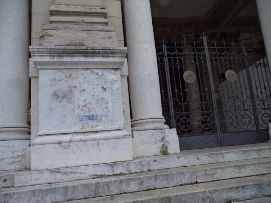 Outside a historic building in Messina. Three marble steps lead up to decorative wrought iron gates. The marble bases of two Tuscan columns can be seen on the left side.