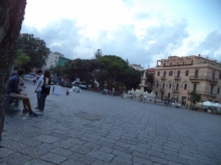 View across Piazza Duomo which is located in front of Messina Cathedral. The square is paved with rectangular stone blocks. A few people can be seen standing or sitting on benches. At the far side a large ornate marble fountain can be seen. This is the Fountain of Orion (Fontana di Orione) designed by Giovanni Angelo Montorsoli and dating from 1553. The base of the fountain is 12-sided and includes four statues depicting the rivers Nile, Tiber, Ebro, and Camaro. In the centre there is an ornately decorated column incorporating several statues. The column supports two raised basins. At the top there is a statue of Orion with his dog Sirius at his feet.