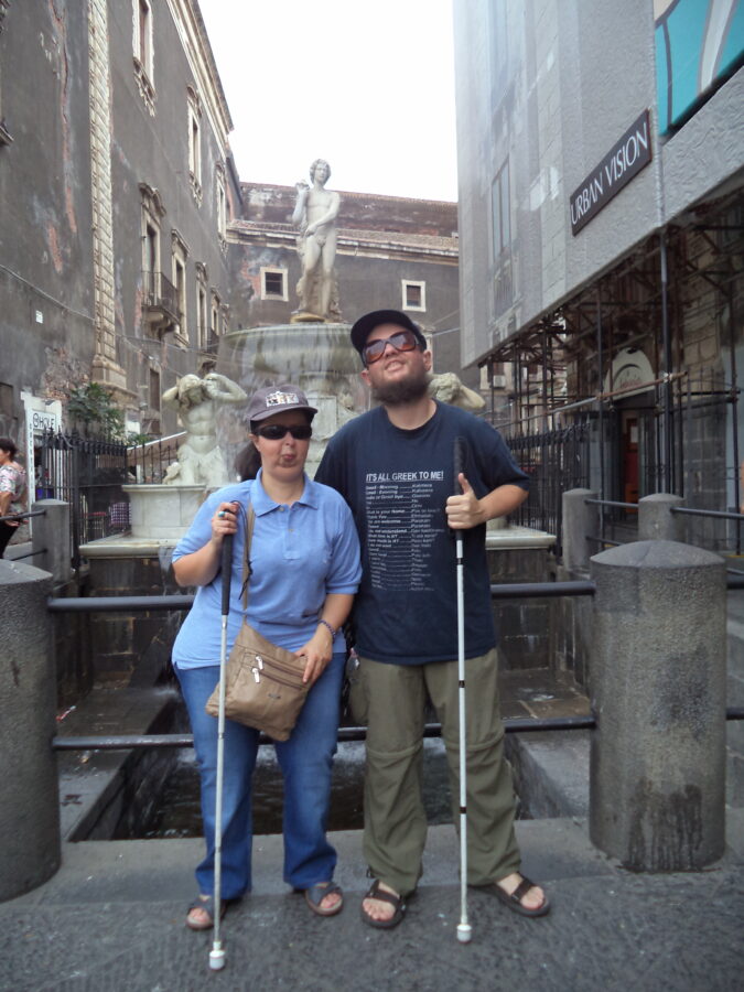 Tony and Tatiana in central Catania standing in front of the Amenano Fountain (Fontana dell'Amenano). This attractive fountain was carved in 1867 by the Neapolitan master Tito Angelini in Carrara marble. The fountain is named after the Amenano river which flows underground beneath Catania. On the top layer of the fountain stands a carved male figure: he personifies the Amenano river and holds a container from which river water flows. The water then flows into a larger pool at the base of the fountain. Two more male statues stand on the base and they depict Tritons or sea gods.