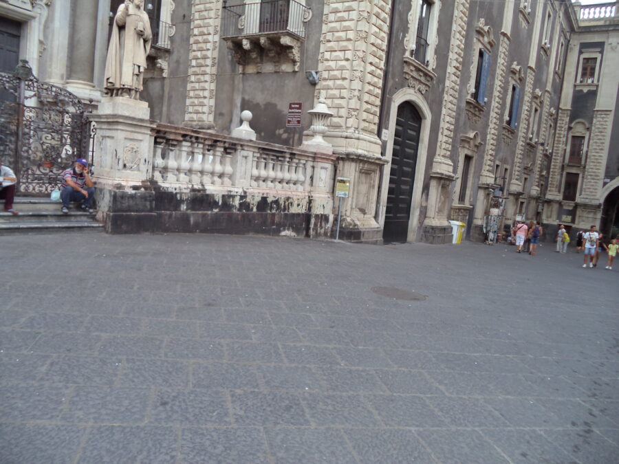The south-east side of stone-paved Piazza dell Duomo. Grand historic buildings can be seen. To the left, a small part of the cathedral is in view. A pale stone balustrade wall stands in front with a statue, likely of a saint, mounted on a large stone gate post at the near end. It has an ornate metal gate attached. The large Baroque building on the right is part of the Palazzo del Seminario dei Chierici (Palace of the Seminary of the Clerics). This wing of the building today houses the Museum of Catania Diocese.  The arch just visible further to the right is the 18th-century Uzeda Gate (Porta Uzeda). This provides a southern exit from Piazza dell Duomo.