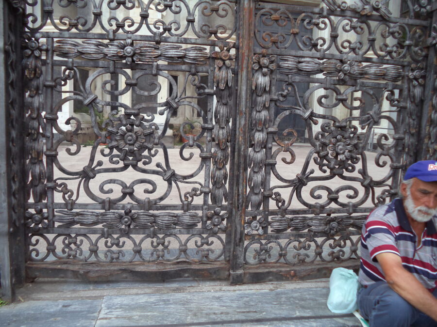 Ornate wrought iron gates with intricate floral and geometric patterns. This is one of several gates outside Catania Cathedral. An elderly man with a white beard and a blue cap is sitting on a step at the bottom.