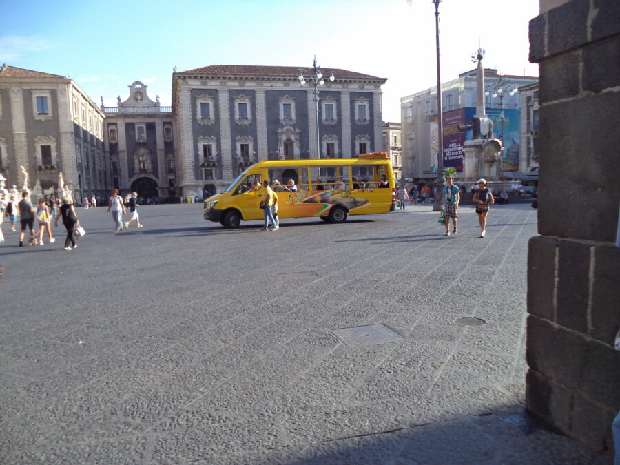 Another view across busy Piazza dell Duomo towards the historic Palazzo del Seminario dei Chierici (Palace of the Seminary of the Clerics) on the south side. A yellow open-sided tourist minibus is parked in front. The Elephant Fountain (Fontana dell'Elefante) is again visible to the right.