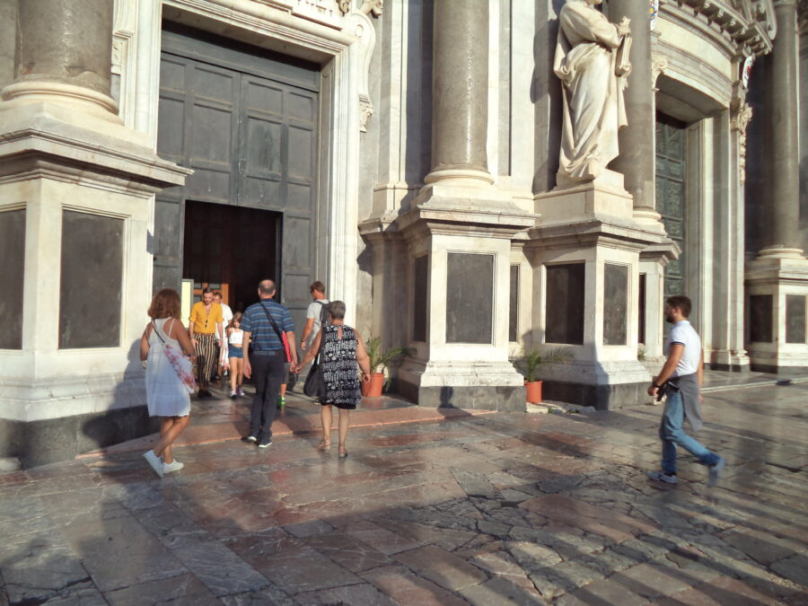 Large doorway into Catania Cathedral with several visitors entering and exiting the building. The door has a rectangular profile with a classical-style stone surround. There are large marble columns rising at the sides. There is also a large marble statue on a pedestal. It depicts a man dressed in robes and holding a book (unfortunately his head is out of view). This might be Saint Birillus. To the right, part of the larger central door, can also be seen.
