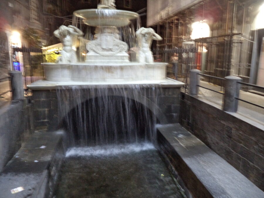 Good view of the Amenano Fountain (Fontana dell'Amenano) taken during the evening. The picture shows the lower part of the fountain. Two male figures carved in marble are depicted in a crouched position, they are in classical style, and represent Tritons or sea gods. Water flows over these figures from an upper bowl down into a larger bowl at the base and from here the water flows below street level into a channel along which the Amenano river flows. Today the Amenano river mostly flows underground, but a short section is visible here. The river (in size more like a stream) emerges from a stone arch.