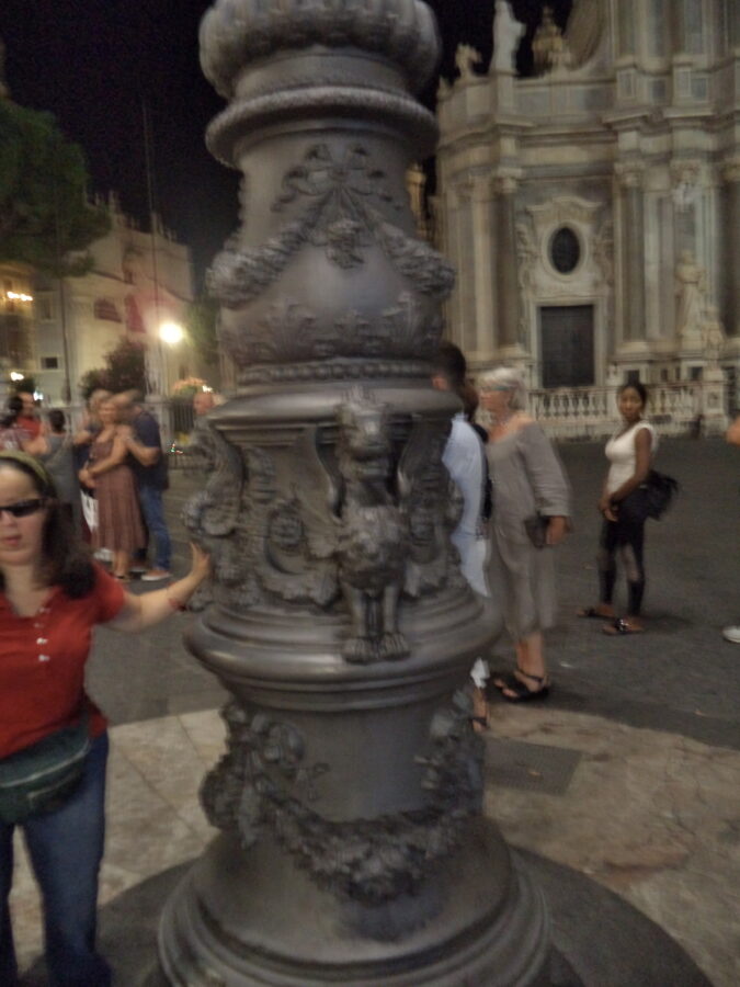 Tatiana stood at the base of a large lamp post located in Piazza dell Duomo during the evening. The pedestal of the lamp post is elaborately decorated with floral motifs and also dragons. One of these metal dragons can prominently be seen looking towards the camera with its wings open at the sides. The square is busy with people in the background.